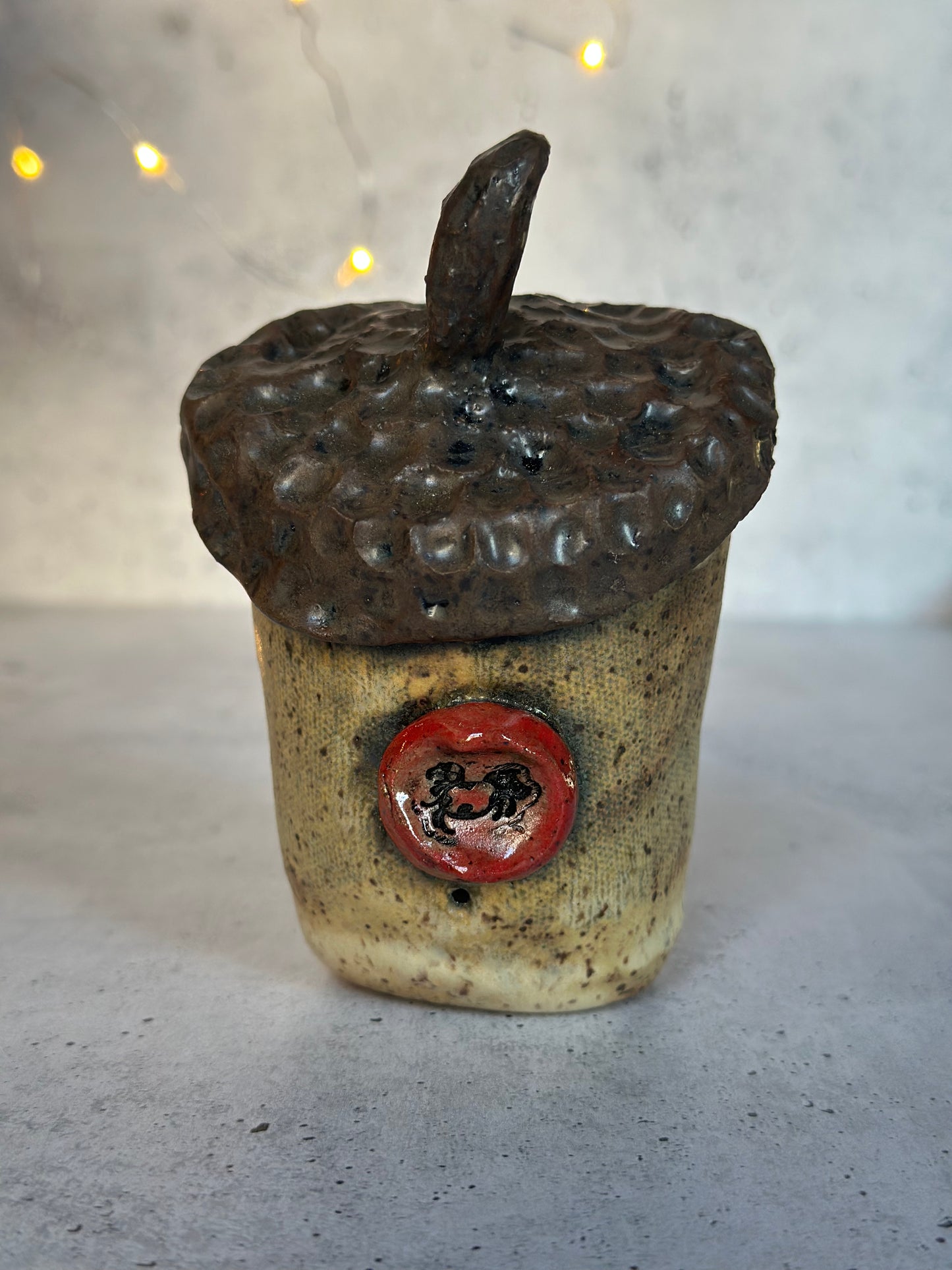 Ceramic acorn-shaped container with a red emblem on a gray surface