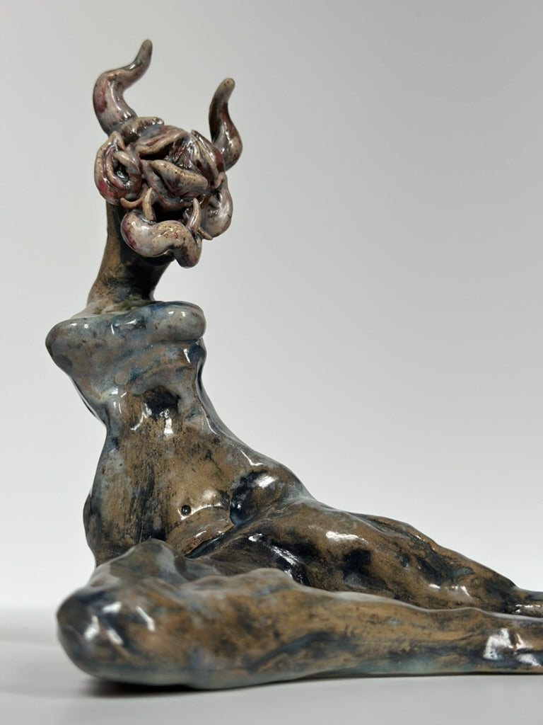 A sculptural figurine of a reclining female form with curled horns on the head, shown in glossy dark gray, brown, and blue-green glaze tones against a light gray studio background.