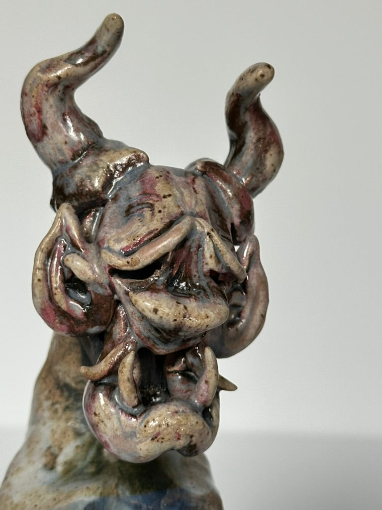 A close-up of a ceramic sculpture depicting a horned, snarling beast or demon head, featuring a textured glaze with brown, gray, and muted blue tones against a neutral light gray background.