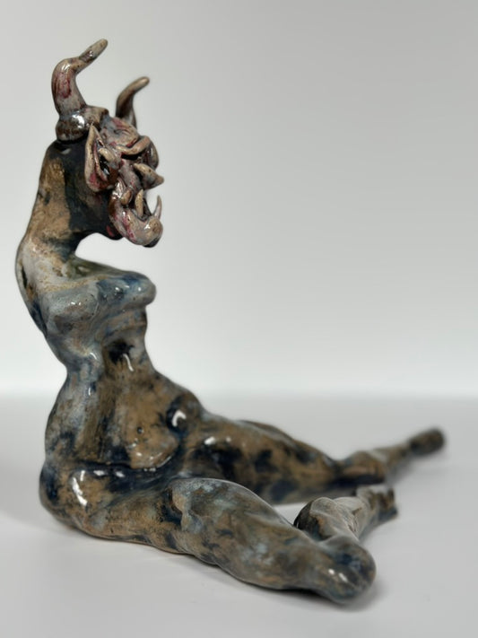 A small sculptural figurine of a seated horned demon or beast, shown on a plain light gray studio background. The object has a glossy glaze with mottled dark blue, gray, brown, and coppery metallic tones.