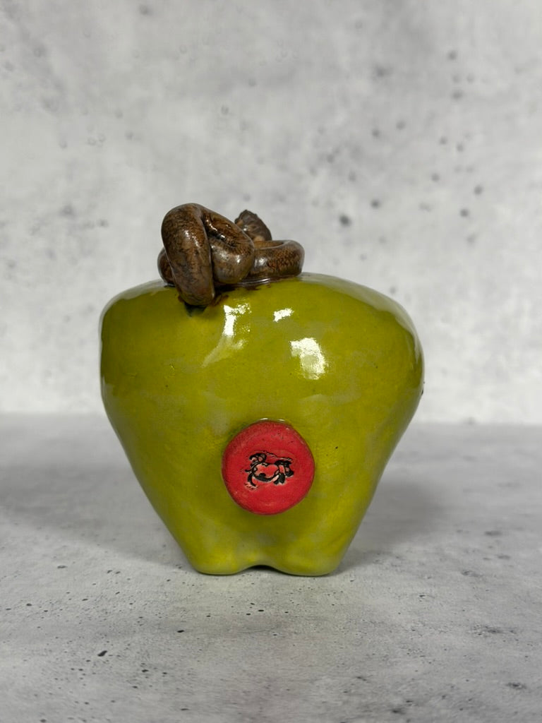 Green ceramic apple-shaped object with a red label and brown knot on a gray background