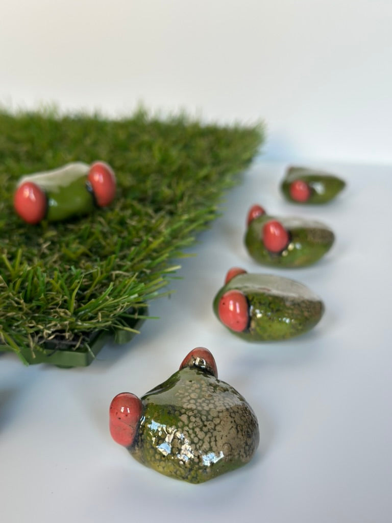 Set of ceramic frogs with red eyes on a white surface with greenery.