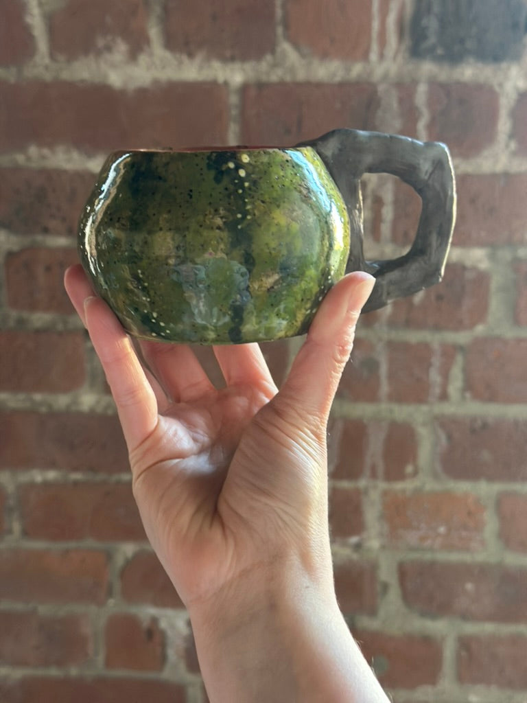 Hand holding a green ceramic mug against a brick wall