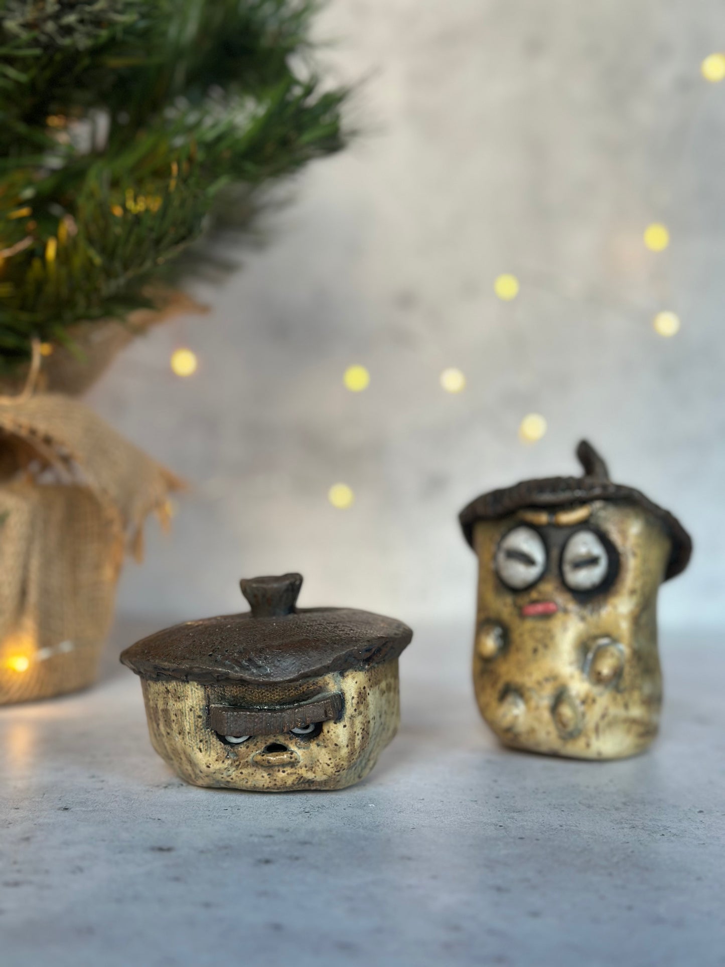 Two small ceramic acorns with faces and hats on a neutral background with a Christmas tree.