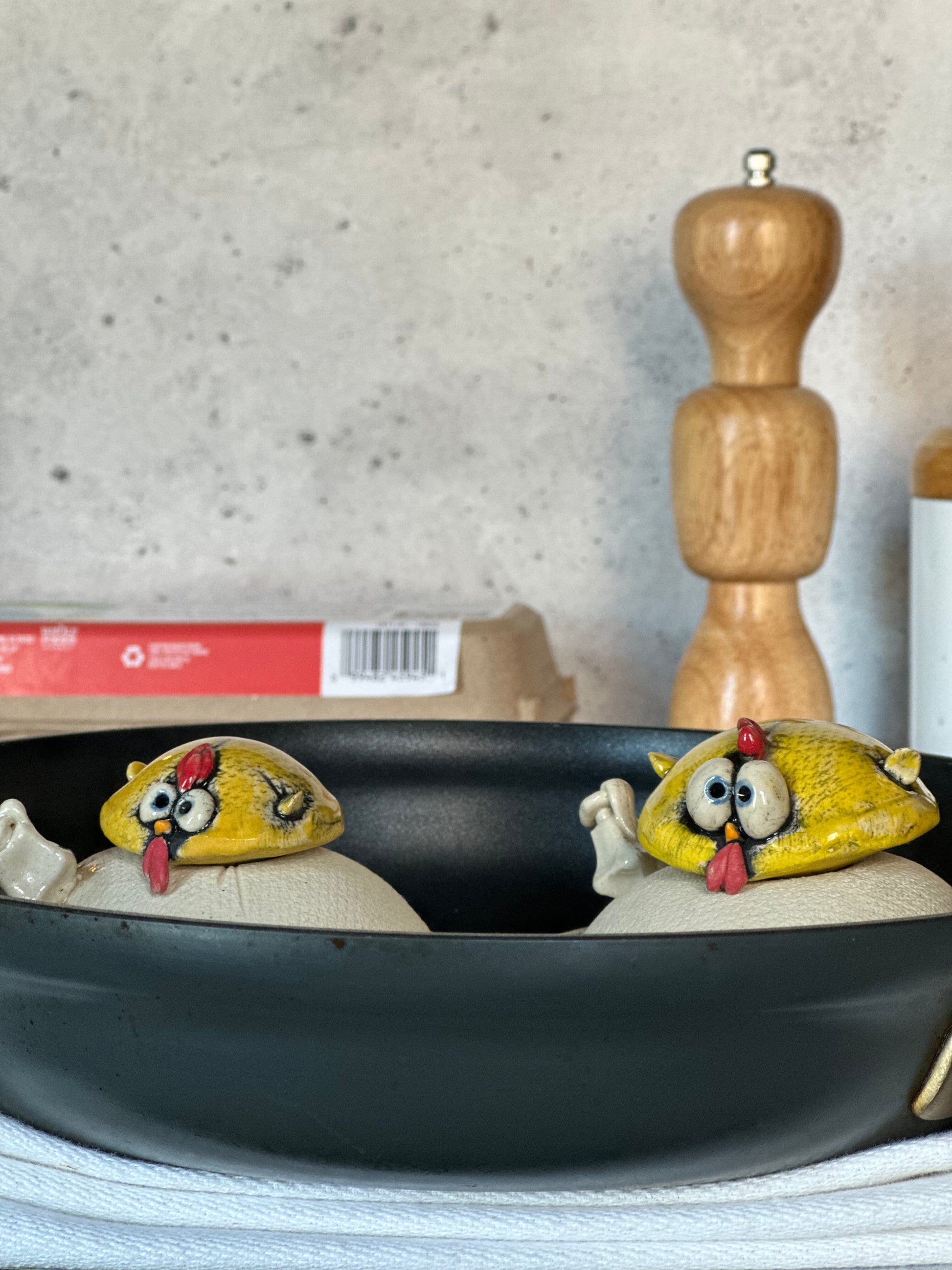 Two cartoonish chicken-shaped objects in a frying pan with a wooden pepper grinder in the background.