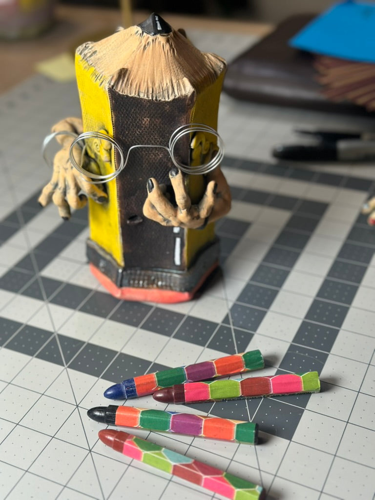 A novelty pencil-shaped figurine with a face, wire-rimmed glasses, and clawed hands, standing on a cutting mat with several colorful crayons in the foreground.