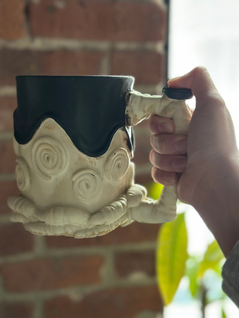 Ceramic mug with a unique design held by a hand against a brick wall.