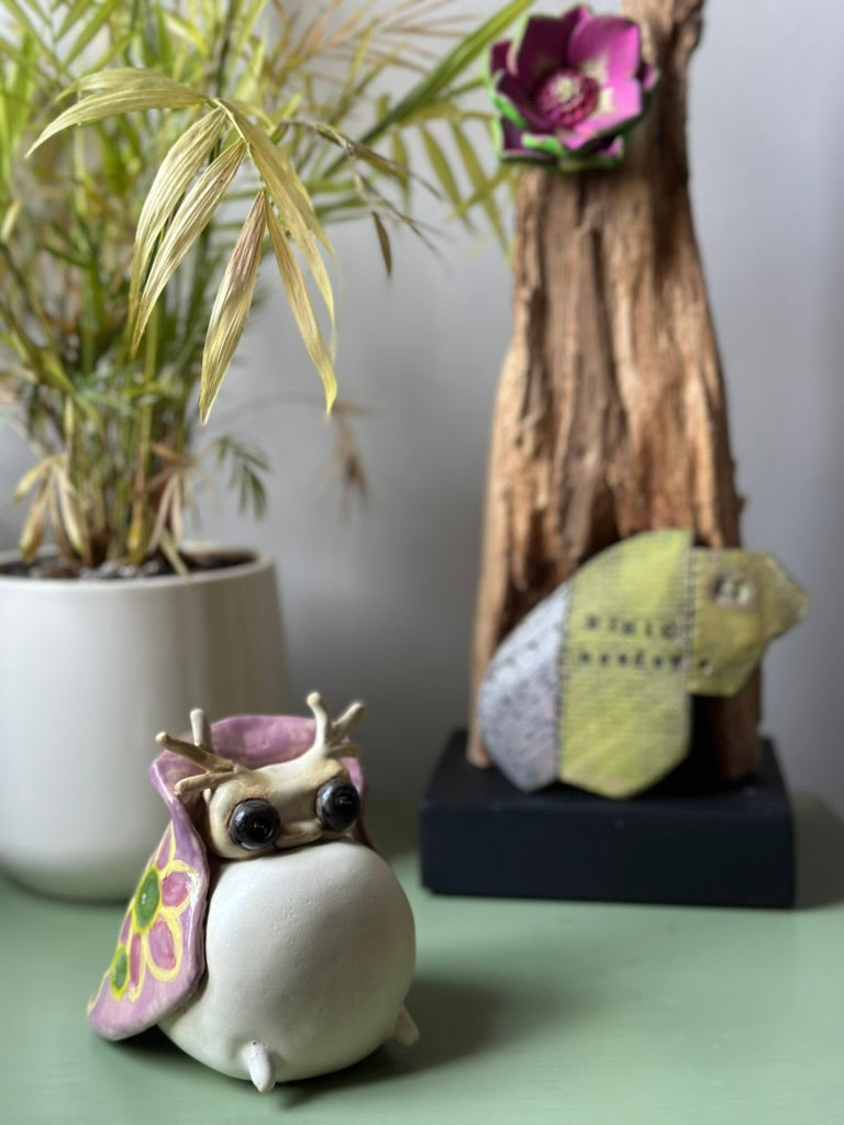Decorative tutrix figurine with colorful wings on a green surface, with a plant and wooden sculpture in the background.