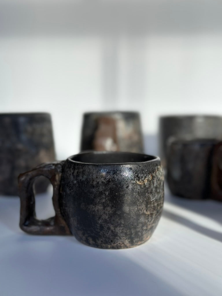Black speckled ceramic mug on a white surface with other mugs in the background